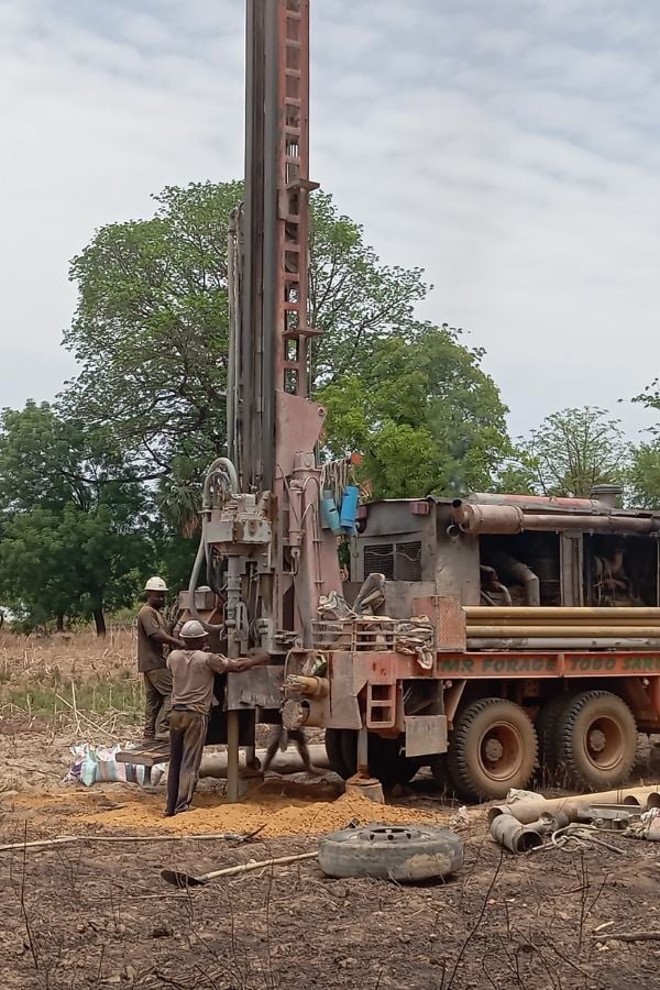 Three workers operate a large drilling rig mounted on a truck in a dry, rural area with trees in the background.