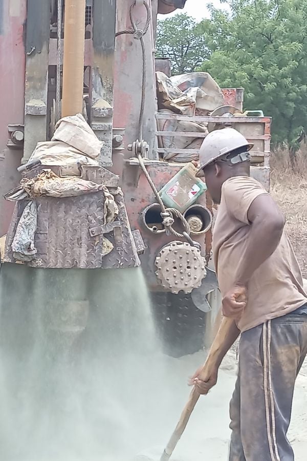 A worker wearing a helmet uses a shovel near heavy machinery as dust pours out from a large pipe in an outdoor, industrial setting.
