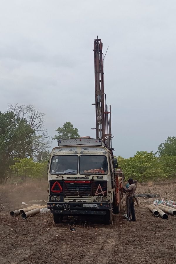 A drilling truck is parked on dry ground with pipes nearby, and two people stand beside it. Trees and cloudy sky are in the background.