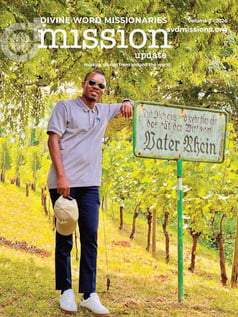 A man in sunglasses leans on a sign by a grassy hillside. The sign reads “Vater Rhein” in German. The photo is the cover of the Divine Word Missionaries magazine.