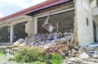 A building with a collapsed wall and debris scattered outside, exposing the interior after apparent structural damage.